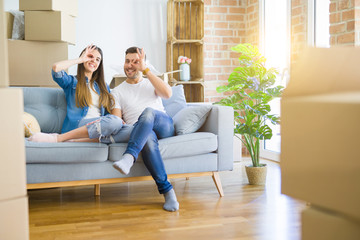 Young beautiful couple relaxing sitting on the sofa around boxes from moving to new house doing ok gesture with hand smiling, eye looking through fingers with happy face.