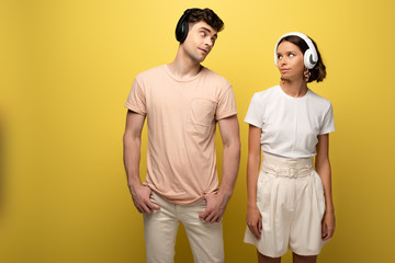 young man and woman listening music in headphones while looking at each other on yellow background