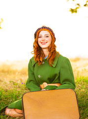 Redhead girl with suitcase sitting at autumn grass