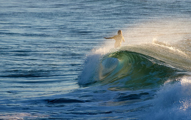 The figure of a female longboarder surfing, partially obscured by the sea-spray from a beautiful breaking-wave; backlit by the early morning sunshine, which is rising over the Pacific Ocean.