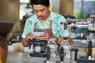 Fototapeta premium Candid lifestyle shot of smiling ethnic indonesian barista and small business owner preparing organic fair-trade coffee in bright trendy coffee shop wearing apron