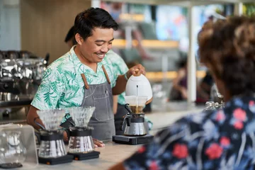 Fotobehang Candid lifestyle shot of smiling ethnic indonesian barista and small business owner preparing organic fair-trade coffee in bright trendy coffee shop wearing apron © AYAimages