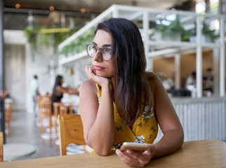 Candid lifestyle portrait of beautiful hispanic millennial woman looking out window holding cellphone in modern trendy and bright coffee shop with plants - 278640755