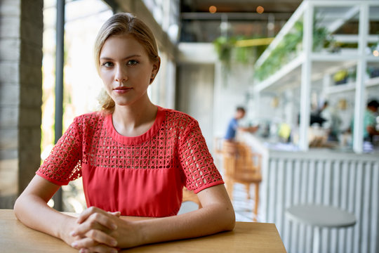 Lifestyle portrait of confident young trendy millennial businesswoman with blonde hair sitting at wooden table in bright and modern restaurant
