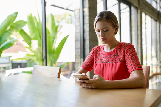 Lifestyle shot of young blonde caucasian natural millennial woman typing on cellphone at a modern tropical and bright office with plants