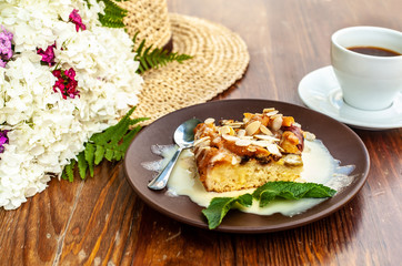 Still life with cake on brown plate, cup of coffee, yellow straw hat and white small flower bouquet on aged wooden table or plate