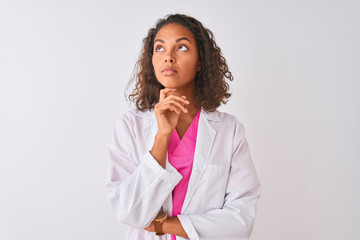 Young brazilian doctor woman wearing coat standing over isolated white background with hand on chin thinking about question, pensive expression. Smiling with thoughtful face. Doubt concept.