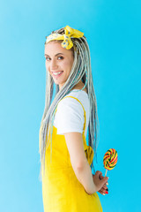 smiling girl with dreadlocks looking at camera while holding lollipop isolated on turquoise