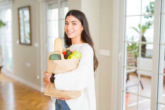 Beautiful young woman smiling holding a paper bag full of fresh groceries at home