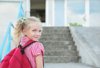 Back to school. Beautiful blond schoolgirl with red backpack outside the primary school from back, education concept.