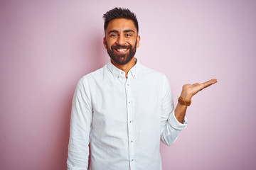 Young indian businessman wearing elegant shirt standing over isolated pink background smiling cheerful presenting and pointing with palm of hand looking at the camera.