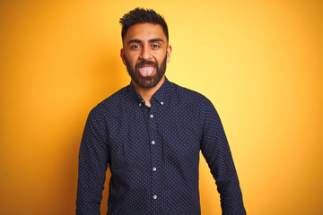Young handsome indian businessman wearing shirt over isolated yellow background sticking tongue out happy with funny expression. Emotion concept.