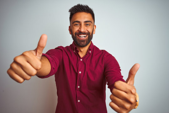 Young indian man wearing red elegant shirt standing over isolated grey background approving doing positive gesture with hand, thumbs up smiling and happy for success. Winner gesture.