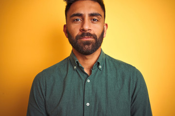 Young indian man wearing green shirt standing over isolated yellow background with serious expression on face. Simple and natural looking at the camera.