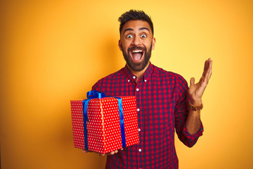Arab indian hispanic man holding birthday gift standing over isolated yellow background very happy and excited, winner expression celebrating victory screaming with big smile and raised hands - Powered by Adobe