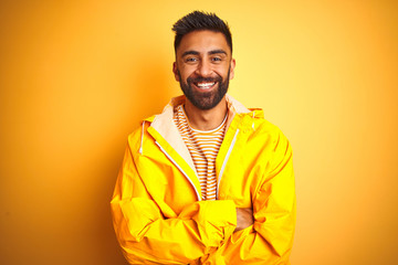 Fototapeta premium Young indian man wearing raincoat standing over isolated yellow background happy face smiling with crossed arms looking at the camera. Positive person.