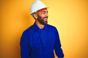 Handsome indian worker man wearing uniform and helmet over isolated yellow background looking away...