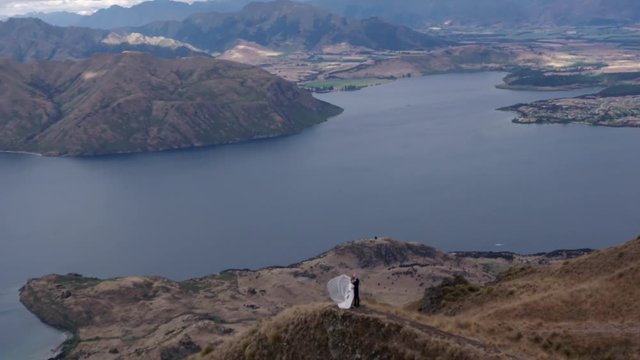Slowmotion helicopter aerial of ust married couple after elopement standing on top of hill of picturesque lake in wedding dress and suit
