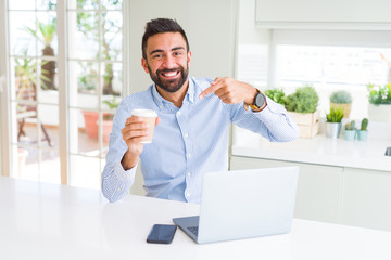 Handsome hispanic man working using computer laptop and drinking a cup of coffee very happy pointing with hand and finger