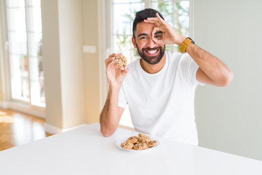 Handsome hispanic man eating chocolate chips cookies with happy face smiling doing ok sign with hand on eye looking through fingers