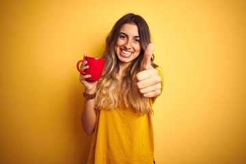 Young beautiful woman holding  red cup of coffee over yellow isolated background happy with big smile doing ok sign, thumb up with fingers, excellent sign