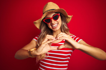 Young beautiful woman wearing sunglasses and summer hat over red isolated background smiling in love showing heart symbol and shape with hands. Romantic concept.