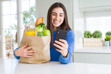 Young woman holding a paper bag full of fresh groceries and using smartphone app for supermarket...