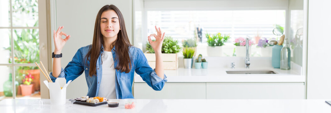 Wide angle picture of beautiful young woman eating delivery sushi relax and smiling with eyes closed doing meditation gesture with fingers. Yoga concept.