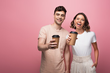 happy man and woman hoding paper cups and smiling at camera on pink background