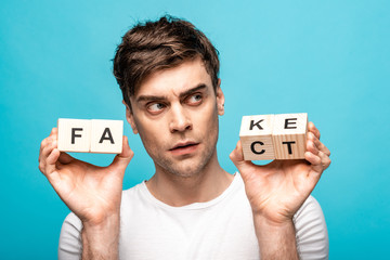 thoughtful young man looking away while holding wooden cubes with fake fact lettering isolated on...