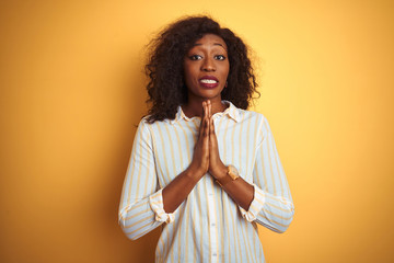 African american woman wearing striped shirt standing over isolated yellow background praying with hands together asking for forgiveness smiling confident.