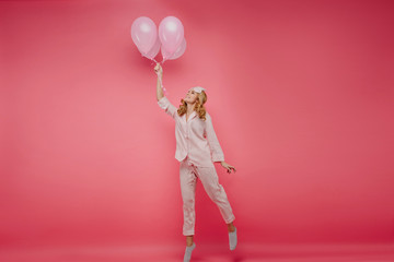 ull-length photo of enthusiastic blonde girl posing with balloons. Indoor portrait of cheerful lady in sleepmask standing on tip-toe in her birthday.