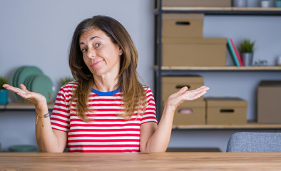 Middle age senior woman sitting at the table at home clueless and confused expression with arms and hands raised. Doubt concept.