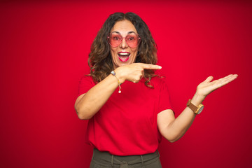 Middle age senior woman wearing cute heart shaped glasses over red isolated background amazed and smiling to the camera while presenting with hand and pointing with finger.