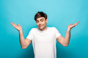 disappointed young man looking at camera and showing shrug gesture on blue background