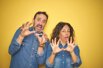 Beautiful middle age couple together standing over isolated yellow background afraid and terrified with fear expression stop gesture with hands, shouting in shock. Panic concept.