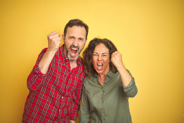 Beautiful middle age couple over isolated yellow background angry and mad raising fist frustrated and furious while shouting with anger. Rage and aggressive concept.