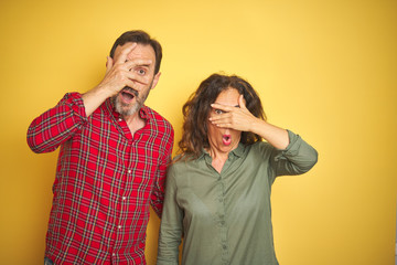 Beautiful middle age couple over isolated yellow background peeking in shock covering face and eyes with hand, looking through fingers with embarrassed expression.