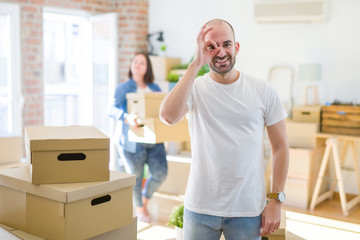 Young couple arround cardboard boxes moving to a new house, bald man standing at home doing ok gesture with hand smiling, eye looking through fingers with happy face.