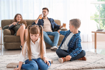 Sad little children sitting on floor while addicted parents drinking alcohol at home