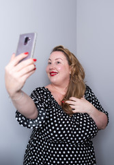 Beautiful overweight plus size model taking selfies for social media with her smart phone. the woman is touching her hair and smiling at camera wearing a black dress with white dots