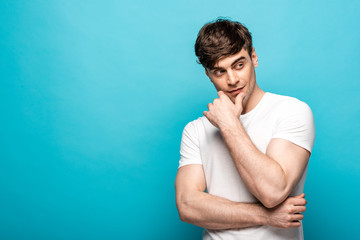 Fototapeta premium pensive young man looking away while holding hand near face on blue background