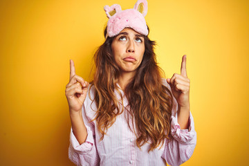 Young woman wearing pajama and sleep mask standing over yellow isolated background Pointing up looking sad and upset, indicating direction with fingers, unhappy and depressed.