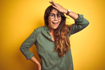 Fototapeta na wymiar Young beautiful woman wearing green shirt and glasses over yelllow isolated background surprised with hand on head for mistake, remember error. Forgot, bad memory concept.