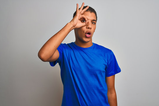 Young handsome arab man wearing blue t-shirt standing over isolated white background doing ok gesture shocked with surprised face, eye looking through fingers. Unbelieving expression.