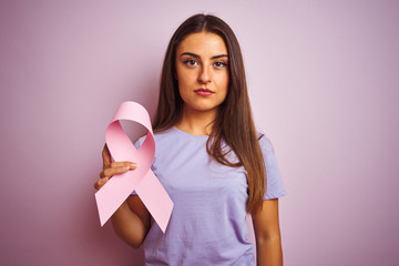 Young beautiful woman holding cancer ribbon standing over isolated pink background with a confident...