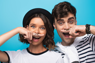 cheerful man and woman holding fingers with drawn mustache near faces on blue background