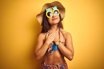Woman on vacation wearing bikini and pineapple sunglasses over isolated yellow background smiling with hands on chest with closed eyes and grateful gesture on face. Health concept.