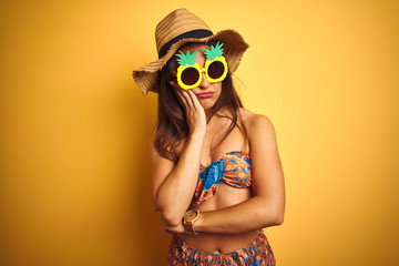 Woman on vacation wearing bikini and pineapple sunglasses over isolated yellow background thinking looking tired and bored with depression problems with crossed arms.