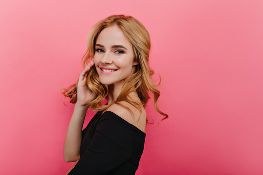Studio shot of blissful woman playing with her blonde hair. Indoor portrait of romantic laughing girl in stylish black dress.